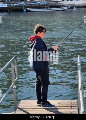 Boy standing on the bank of river Limmat Zurich in Switzerland. He holds a fishing rod. It is launched in water. Side view with water on background. Stock Photo