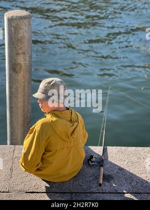 Boy sitting on the bank of river Limmat Zurich in Switzerland. There is a fishing rod beside him. It is launched in water. Rear view with water. Stock Photo