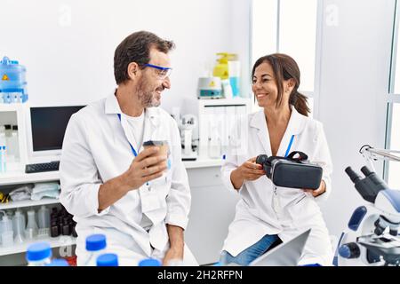 Middle age man and woman partners wearing scientist uniform drinking coffee and holding vr goggles at laboratory Stock Photo