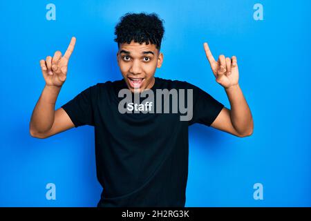 Young african american man wearing staff t shirt smiling amazed and surprised and pointing up with fingers and raised arms. Stock Photo