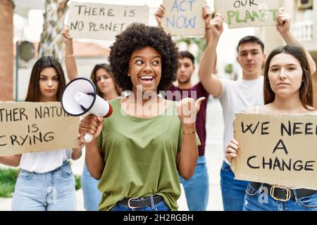 Group of young friends protesting and giving slogans at the street pointing thumb up to the side smiling happy with open mouth Stock Photo