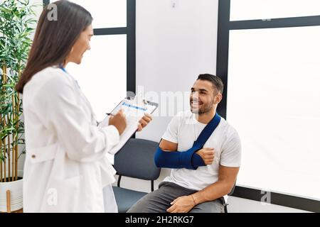 Youg latin injuried man wearing sling talking with the doctor at clinic waiting room. Stock Photo