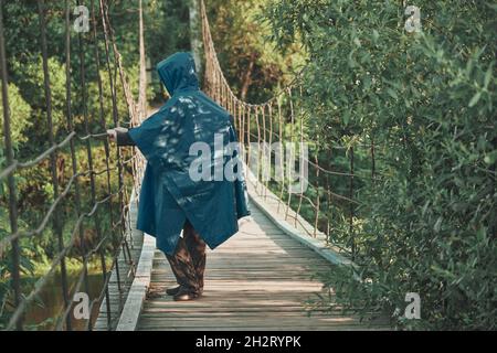 Girl with backpack under blue raincoat stands on suspended, wooden bridge Stock Photo
