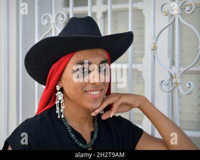 Handsome young nonbinary Latino with black hat, red Mexican rebozo and Day of the Dead skeleton earrings smiles in front of white window grill. Stock Photo