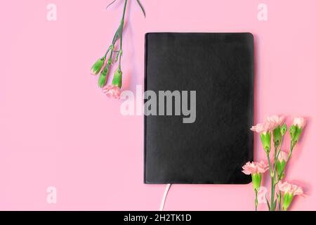 Top view of black empty book or notebook and flowers on pastel pink background with copy space. Stock Photo
