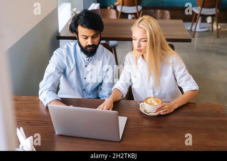 High-angle view of two serious male and female business colleagues sitting at table and talking in cafe looking at laptop screen. Stock Photo