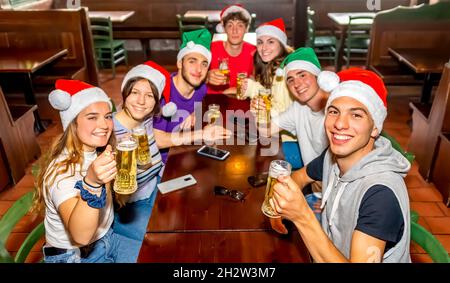 merry christmas! Young group of teenagers friends portrait celebrating xmas time wearing santa claus hat on happy hour in a irish pub drinking beers Stock Photo