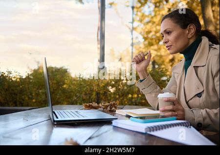 Beautiful business woman concentrated on distant work, looks at the screen of laptop and enjoys outdoor coffee break during lockdown in the terrace of Stock Photo