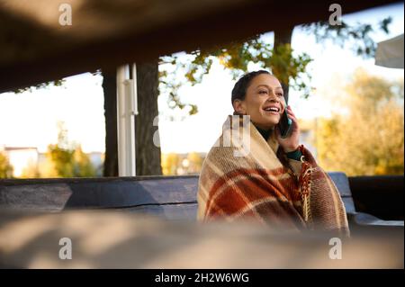 View through wooden logs of bench at smiling pretty woman looking away while talking on cell phone, sitting on wooden bench wrapped in cozy checkered Stock Photo