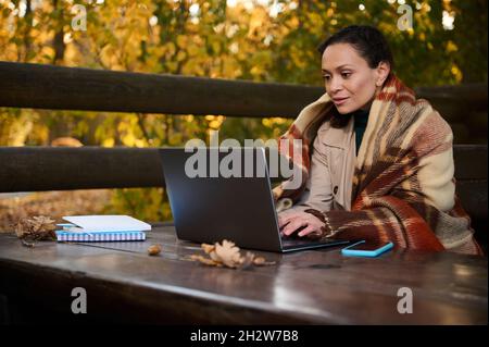 Middle aged freelancer woman, developer wrapped in warm woolen blanket, types on laptop keyboard, works project, plans startup, sits at wooden table i Stock Photo