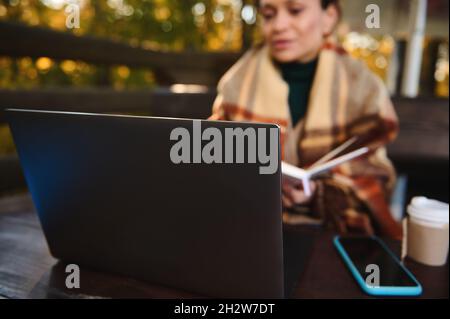 Focus on open laptop on wooden table near mobile phone against the background of blurred woman wrapping herself in a cozy plaid blanket, keeping herse Stock Photo