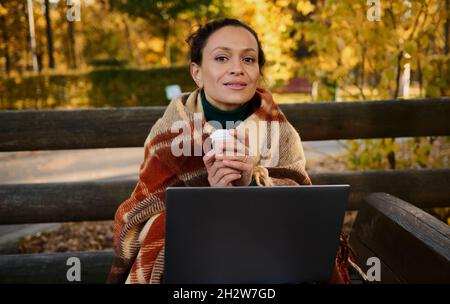 Charming self-confident young woman keeps herself warm wrapped in cozy warm woolen plaid, holds paper cup with hot drink sits on a wooden bench in fro Stock Photo
