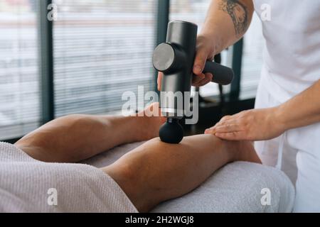 Close-up of unrecognizable professional male masseur massaging leg calf muscles using massage gun percussion tool of muscular athlete man, Stock Photo