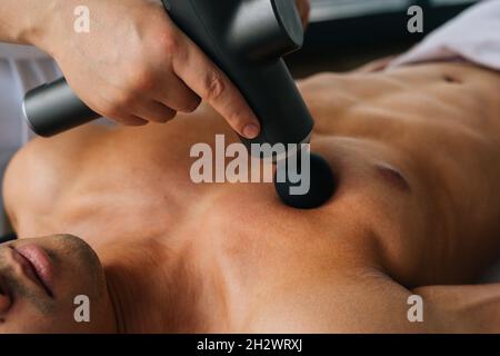 Close-up of unrecognizable professional male masseur receiving chest muscles using massage gun percussion tool of muscular athlete man Stock Photo
