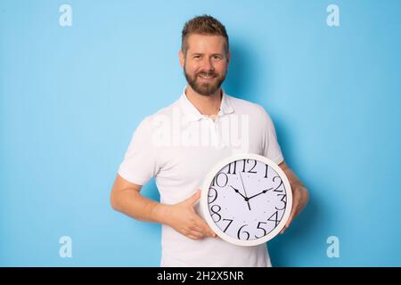 Young man with beard holding big clock over isolated blue background happy with big smile. Stock Photo