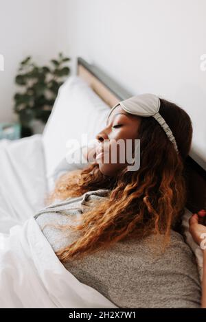 A beautiful young black female laying on the bed with a sleeping mask stretching after waking up Stock Photo