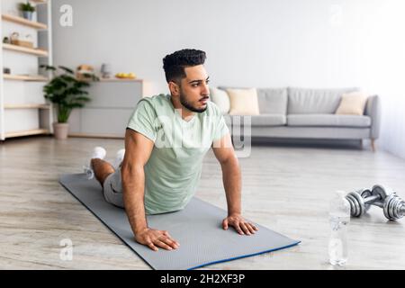 Domestic yoga practice. Flexible young Arab man doing cobra pose on mat, making exercises at home, free space Stock Photo