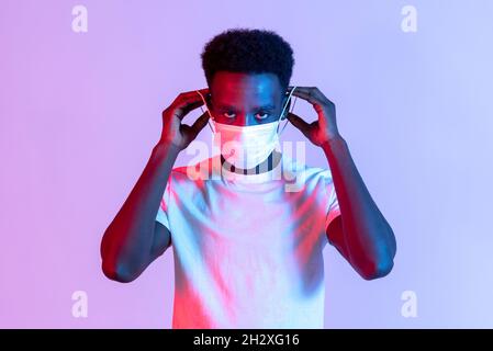 Young Black man preparing to put on a medical face mask during the Covid-19 pandemic to protect against the virus stretching the elastic to place it o Stock Photo