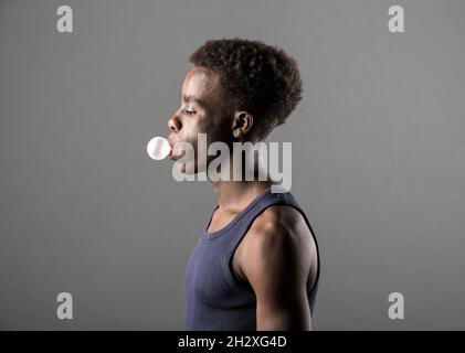 Side view of a trendy sporty young Black man in T-shirt blowing bubbles with chewing gum over a grey studio background in a lifestyle portrait Stock Photo