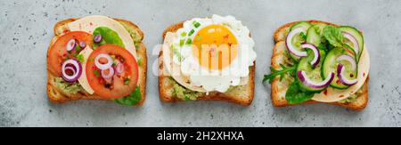 Three different toast sandwiches with guacamole sauce, fried egg, fresh cucumber, cheese and tomato with chard, arugula, red onion on gray concrete ba Stock Photo