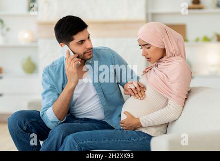 Pregnant arab woman having prenatal contractions, worried husband calling doctor, using mobile phone, home interior Stock Photo