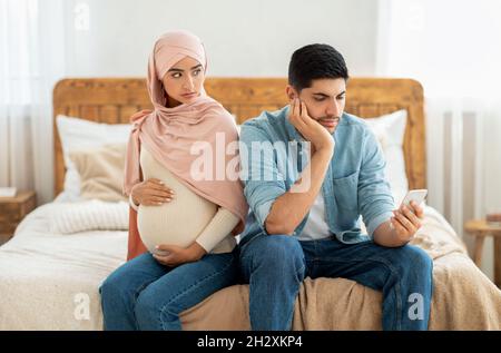 Young pregnant muslim woman offended to her husband busy with smartphone, arab lady in hijab feeling lonely Stock Photo