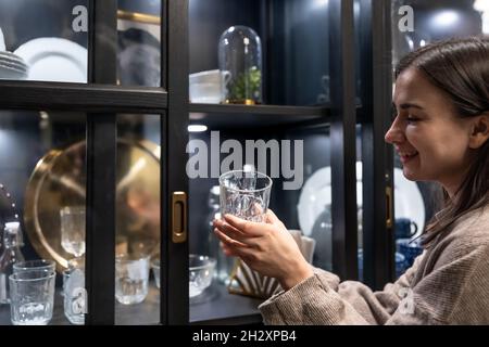A woman holds a glass cup on the background of a cabinet with utensils. Stock Photo