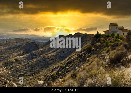 Sunset over the mountains and the house, on top of the mountain, illuminated by the orange light of the hiding sun.Spain, Alicante province.Horizontal Stock Photo