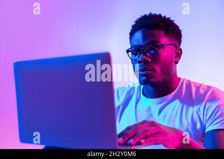 Focused young black man in glasses using laptop computer for online work or communication in neon light Stock Photo