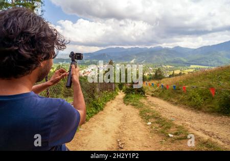 Back or rear view of a male hiker in casual clothes stands in a mountain trail with a sport camera on a stabilizer gimbal and makes a video. Video ope Stock Photo