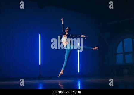 Young woman in casual style - jeans and leather jacket doing ballet in modern studio. Attractive ballerina practices in choreography alone Stock Photo