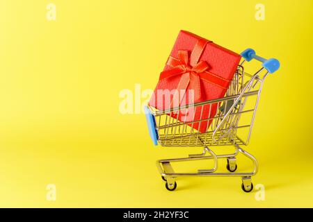 Mini cart with small red gift box on yellow background. Shopping concept in stores and on the internet. The concept of online shopping for the holidays. A universal gift for all holidays Stock Photo