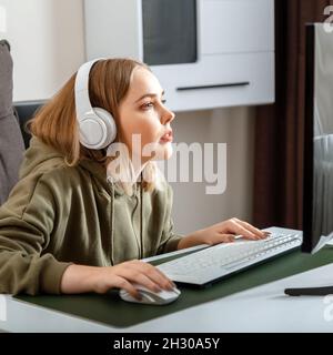 Teenager girl cybersportman gamer play online computer game at home alone in leisure time. Young blonde woman in headphones enthusiastically play pc Stock Photo