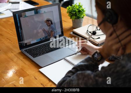 Young student watching lesson online and studying from home. Young woman taking notes while looking at computer screen following professor doing math Stock Photo