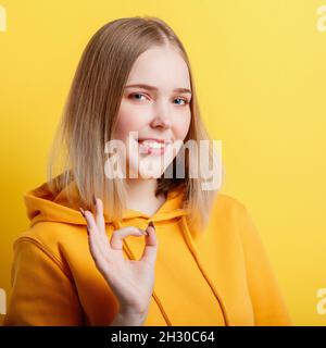 Teenage blonde girl smiles and shows positive ok gesture symbol like and approval isolated on color yellow background. Portrait young woman shows ok Stock Photo