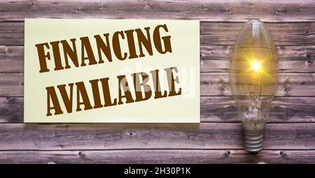 Funding is available written on a white sheet, next to a light bulb. Wood background Stock Photo