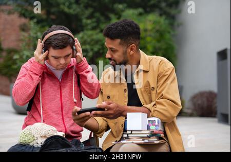 Young man with Down syndrome with his mentoring friend sitting outdoors and listening to music. Stock Photo