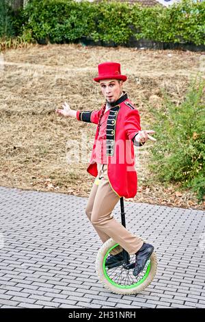 Full body man in costume and hat stretching out arms and balancing while riding unicycle on pavement during performance in park Stock Photo