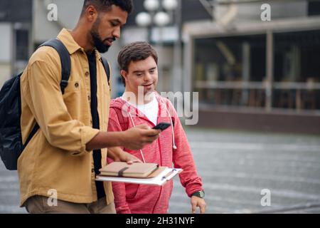 Young man with Down syndrome with his mentoring friend with smartphone walking and talking outdoors Stock Photo