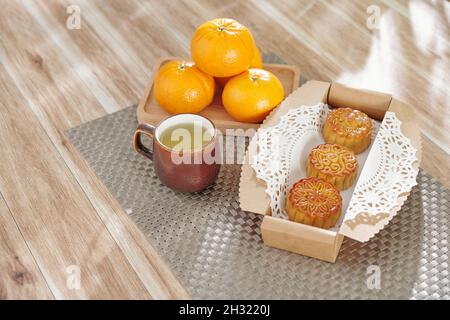 Box of glazed moon cakes, cup of herbal tea and plate with ripe tangerines on kitchen table Stock Photo