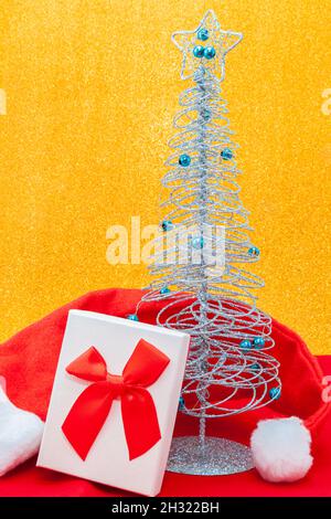 A gift box with a bow next to a glitter Christmas tree with blue beads, surrounded by a Santa Claus hat on a red fabric with a shiny gold background. Stock Photo