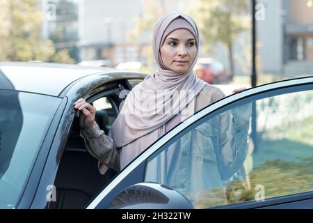 Young woman in hijab standing by open door of her car while getting out of it in urban environment Stock Photo