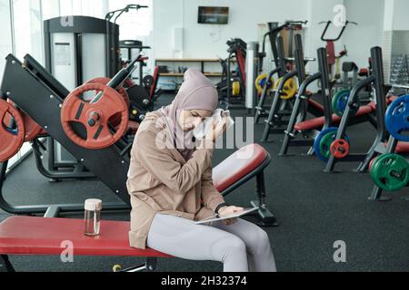 Young restful sportswoman touching her wet face by towel while sitting on exercise equipment after training Stock Photo