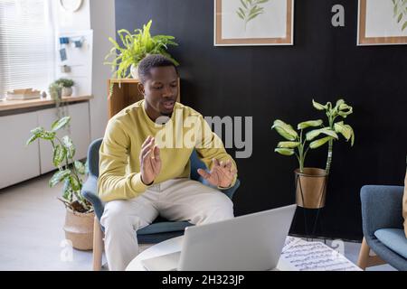 Young confident African-American music teacher sitting in armchair in front of laptop during online lesson Stock Photo