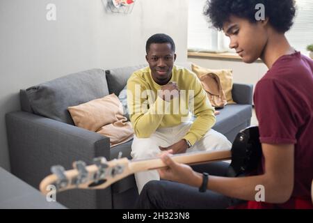 Happy blackman in casualwear looking at young guy playing guitar during music lesson in home environment Stock Photo