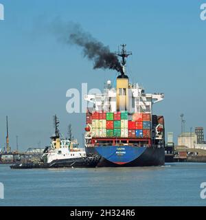Arriving cargo container freighter ship & black smoke stacked with load shipping containers tugboat pushes towards cranes Felixstowe port Suffolk UK Stock Photo