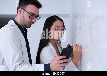 Two researchers in lab coat writing notes on whiteboard. Stock Photo