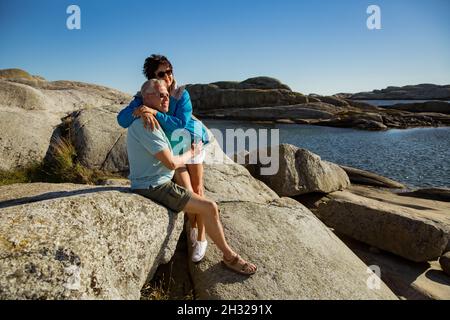 Loving mature couple traveling, sitting on the rock, exploring. Real man and woman hugging, kissing, Happily smiling. Scandinavian landscape, Norway Stock Photo