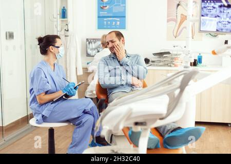 Medical nurse examining patient in paing writing dental problem on clipboard while sitting in dental hospital office. Sick man waiting for orthodontic consultation discussing medical treatment Stock Photo