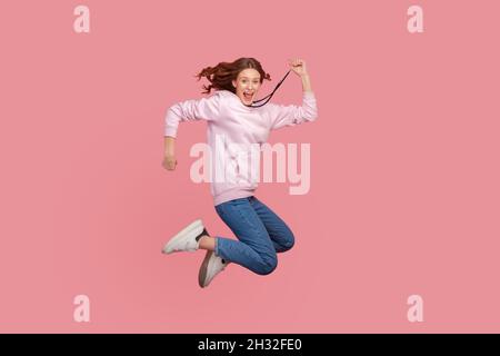 Full length portrait of extremely happy teen girl with curly hair in hoodie jumping for joy or flying, celebrating success, victory, Indoor studio shot isolated on pink background Stock Photo